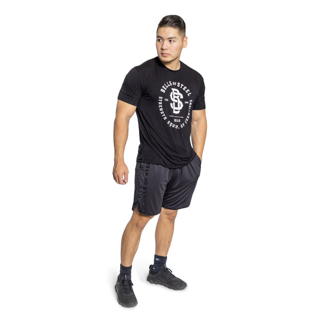 A person stands against a white background wearing Bells of Steel black gym shorts and a T-shirt with a circular design, one hand in their pocket. With black socks and athletic shoes, they look to the side, exuding casual confidence.