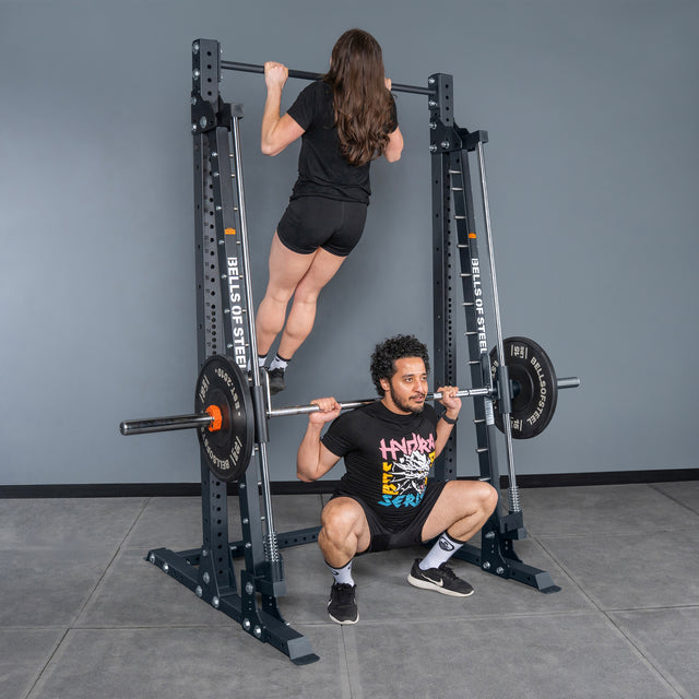 A woman does a pull-up while a man squats with a barbell under the Bells of Steel Smith Machine - Hydra (3" x 3", ⅝" Holes) in a gym with gray walls and floor tiles.