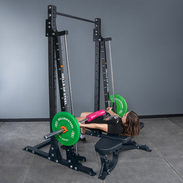 A person performs a hip thrust on a Bells of Steel Smith Machine - Hydra (3" x 3", ⅝" Holes), supporting a barbell with green plates and a pink pad across their hips in a modern gym.