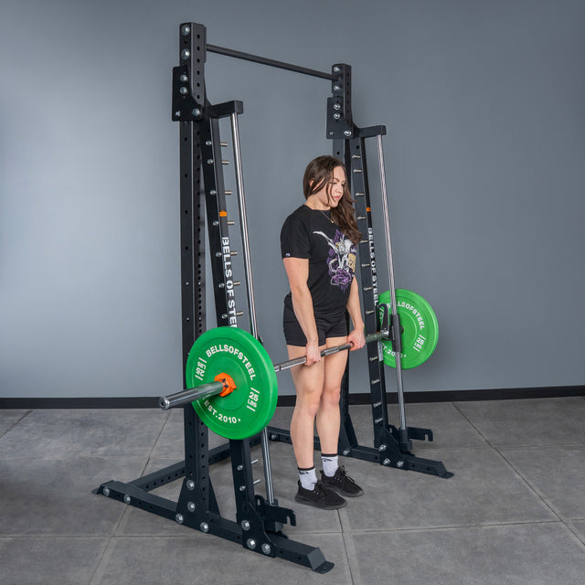 A person stands on a gym floor doing a deadlift with green plates and a barbell, inside the Bells of Steel Smith Machine - Hydra (3" x 3", ⅝" Holes), which features an attached pull-up bar, set against a gray wall.