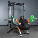 Wearing black and a cap, a person performs a barbell squat with green plates on the Bells of Steel All-in-One Trainer squat rack in a gym with gray concrete floors and walls.