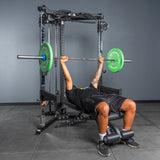 A person bench presses a barbell with green weight plates inside the Bells of Steel All-in-One Trainer in a gym with gray walls and flooring.