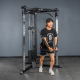 Wearing athletic gear, a person uses the Bells of Steel All-in-One Trainer at the gym, pulling cables downward and forward while standing staggered on a gray floor.