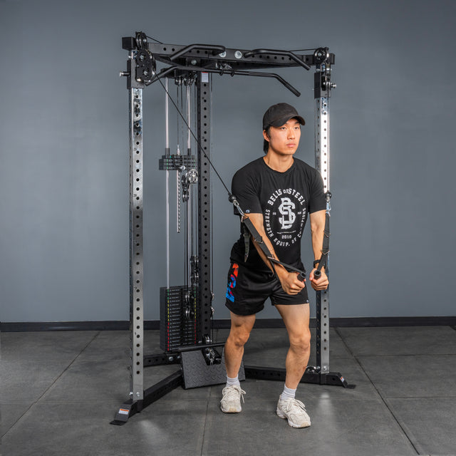 Wearing athletic gear, a person uses the Bells of Steel All-in-One Trainer at the gym, pulling cables downward and forward while standing staggered on a gray floor.