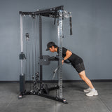 Wearing black athletic gear and a cap, a person performs a triceps pushdown on the Bells of Steel All-in-One Trainer in the gym, leaning forward with arms extended and feet staggered on the gray floor.