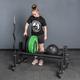 A person in athletic wear loads weight plates onto the Bells of Steel Rack Junctions - 3" x 3", with a barbell and weighted ball nearby on a gray gym floor—ideal for a versatile home gym setup against a plain gray wall.