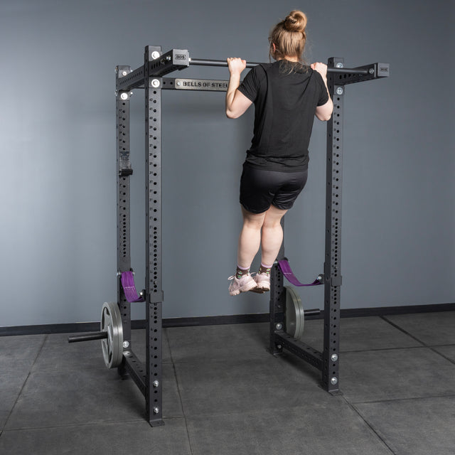 A person with a bun hairstyle does a pull-up on a customizable rack featuring Bells of Steel Rack Junctions - 3" x 3" in a gym, with weight plates attached to the junctions and gray mats covering the floor.