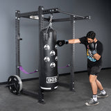 Wearing black boxing gloves, a person punches a black bag in a gym with gray walls, weights, and the Bells of Steel Rack Junctions - 3" x 3", an ideal customizable rack for any home gym setup.