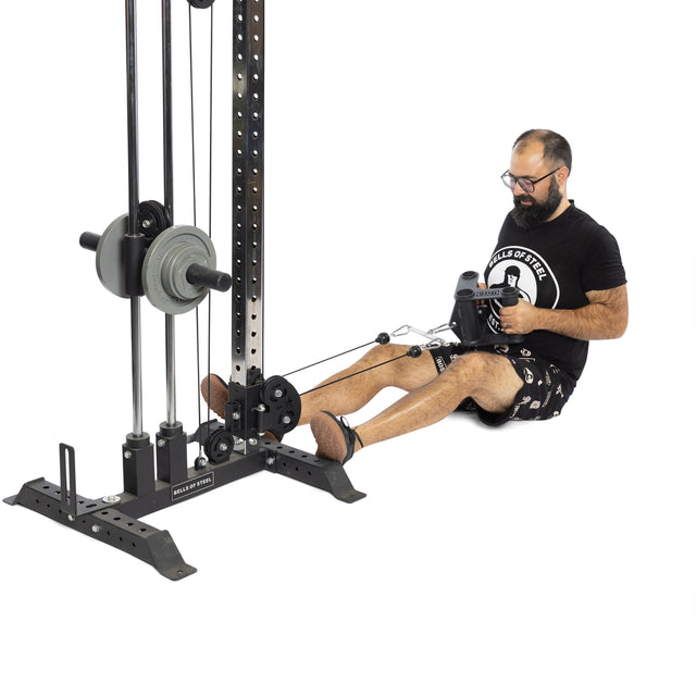 A bearded man in a black t-shirt and shorts uses the Bells of Steel Fat Bar - T-Bar Row Cable Attachment with weight plates for grip strength, seated on an exercise machine.