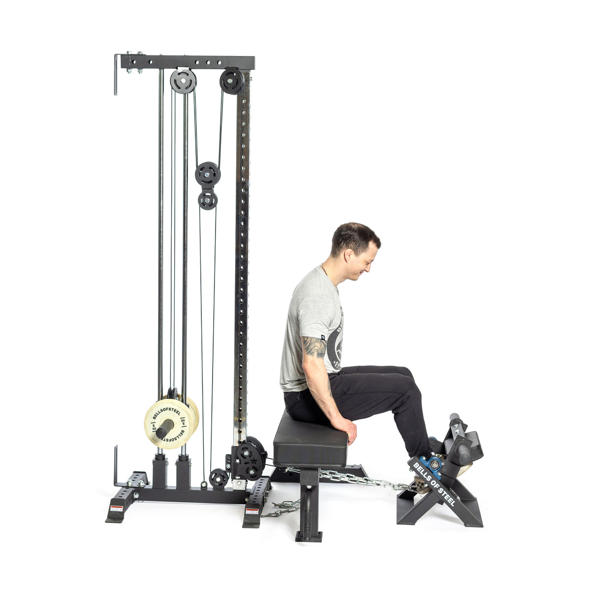 A person seated on a bench is performing a calf raise using the Tibialis Trainer Machine by Bells of Steel, lifting a chain-weighted barbell with their feet while maintaining a straight back. This workout employs tall machinery with pulleys and weights for optimal hypertrophy.