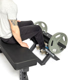 A person sits on a black workout bench, gripping the Tibialis Trainer Machine's edge, with feet on footpads holding two large weights. The "Bells of Steel" brand signifies strength training and hypertrophy against a crisp white background.