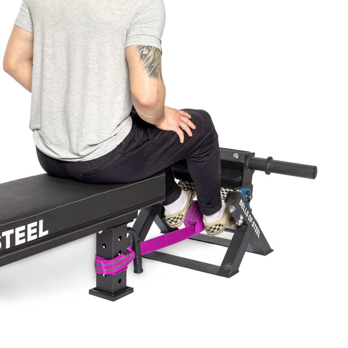 Seated on a black workout bench, a person in a gray t-shirt and black pants, with an upper right arm tattoo, secures their feet with a purple strap to the footrest of the "Bells of Steel" Tibialis Trainer Machine, ideal for enhancing hypertrophy during strength training.