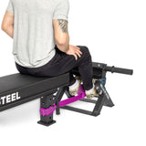 Seated on a black workout bench, a person in a gray t-shirt and black pants, with an upper right arm tattoo, secures their feet with a purple strap to the footrest of the "Bells of Steel" Tibialis Trainer Machine, ideal for enhancing hypertrophy during strength training.