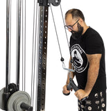 A man in a black shirt and shorts is improving his grip strength by using a cable machine for tricep pushdowns. He is concentrating intently while performing the Fat Bar - Tricep Pressdown from Bells of Steel in a gym setting, gripping the handle securely with both hands.