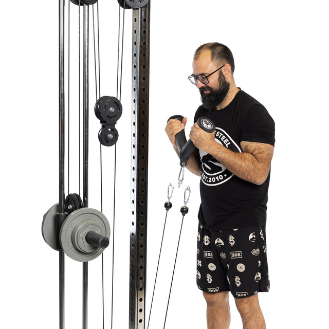 A bearded man in a black t-shirt and patterned shorts is working out on a cable pulley machine, concentrating on tricep pushdowns using the Bells of Steel Fat Bar - Tricep Pressdown. The equipment, equipped with multiple weights and cables, aids him in enhancing his grip strength.