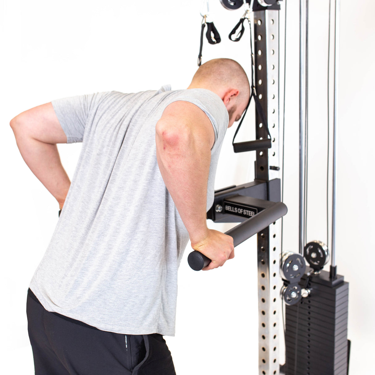 A person in a gray T-shirt and black shorts performs tricep dips on the Bells of Steel Cable Tower, which features adjustable weights and black handles. The machine enhances any home gym, set against a plain white background.