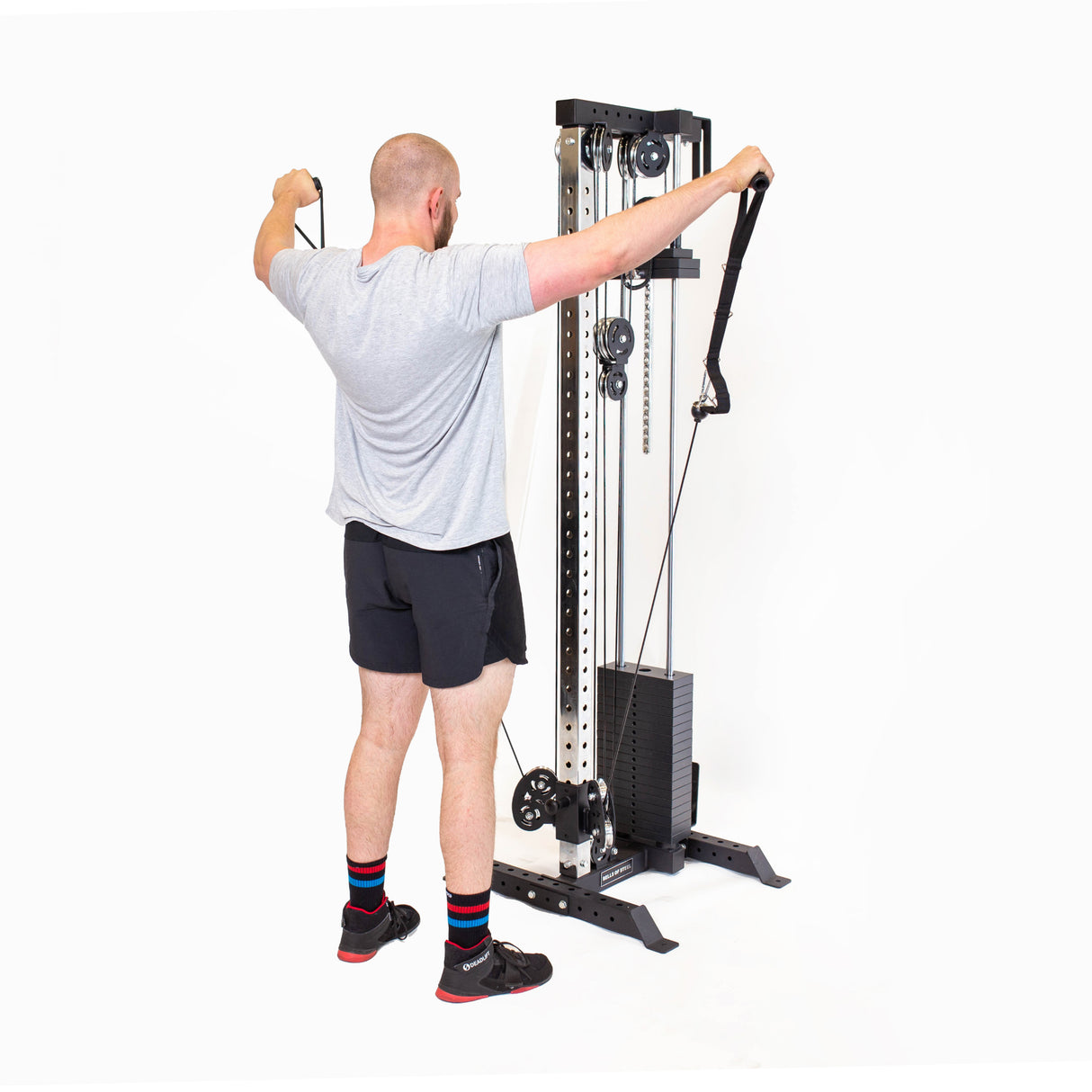 A man in a gray t-shirt and black shorts uses the Bells of Steel Cable Tower for shoulder exercises. He stands with arms outstretched, gripping the handles against a plain white background, evoking a home gym atmosphere.