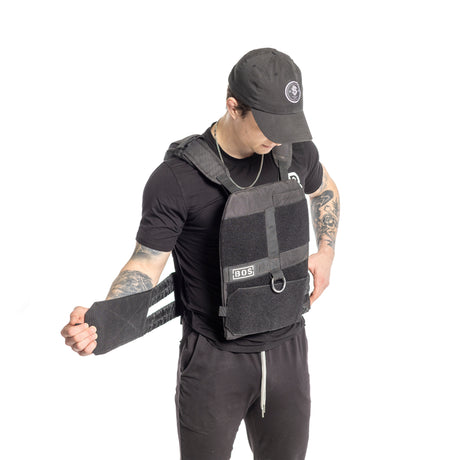 A tattooed person in a black cap and t-shirt adjusts a Bells of Steel Weighted Vest, featuring the logo "BOS." They grip the side strap of this adjustable black tactical vest, set against a white background, ready to add training plates for extra resistance.