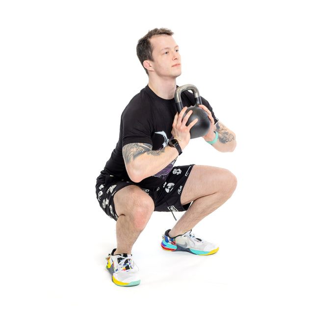 A tattooed man is squatting with a Mark Wildman Adjustable Kettlebell by Bells of Steel close to his chest. He wears a black t-shirt, patterned shorts, and colorful sneakers against a plain white background.