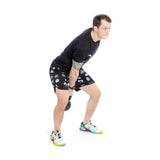 A man in a black T-shirt, patterned shorts, and colorful sneakers uses the Bells of Steel 6-12KG Adjustable Kettlebell for swings, gripping it with both hands between his legs with bent knees and a straight back.