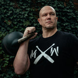 A bald man in a black T-shirt with white crossed bats holds the Bells of Steel Wildman Poison Pill 48KG Extension Kit for Adjustable Kettlebell over his shoulder, standing before a wall of green ivy.