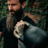 A tattooed, bearded man holds the Bells of Steel 48KG Extension Kit for Adjustable Kettlebell – Wildman Poison Pill over his shoulder, staring intently at the camera; a blurred green and brick wall background completes the scene.
